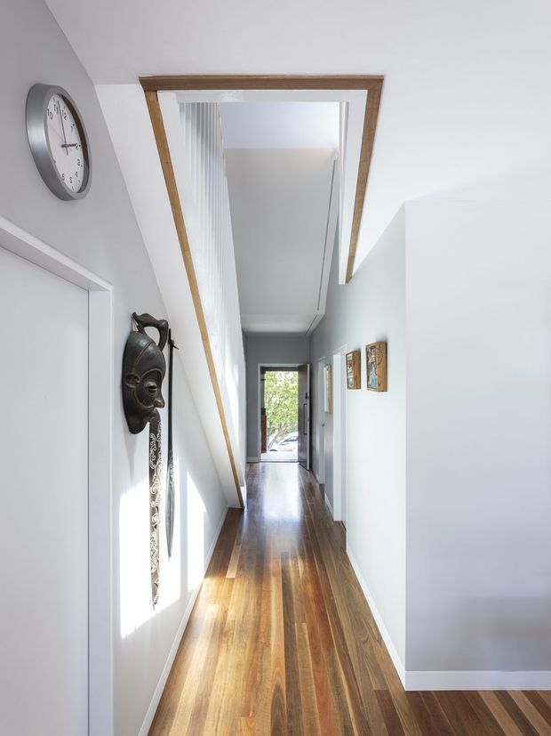 A bright, narrow residential hallway with polished timber flooring, white walls, and a high angular ceiling with exposed timber trim. The corridor features a wall clock, decorative mask, framed artwork, several doorways, and a glazed exit at the far end. Strong natural light creates a clean, airy atmosphere.