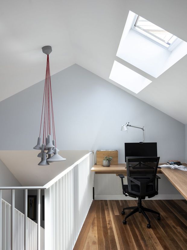 A bright, compact loft-style home office positioned on an upper landing beside a stairwell. The space has white walls and ceiling, two skylights, a timber desk wrapping around the corner, a black computer monitor and ergonomic office chair, and a distinctive cluster pendant light with red cords. White vertical stair balustrades and a dark-to-mid timber floor add architectural definition.