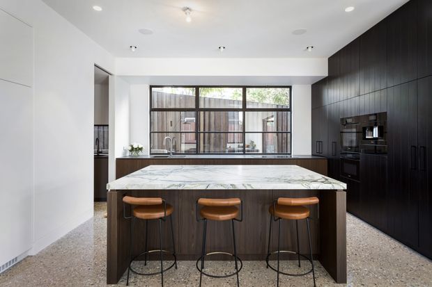 A bright, contemporary kitchen centered on a large marble-look island with seating for three. The room features dark vertical-grain cabinetry and integrated appliances along the right wall, white walls and ceiling, full-height windows above the rear benchtop, and a terrazzo-style floor. Warm leather stools add contrast to the monochrome built-in finishes.