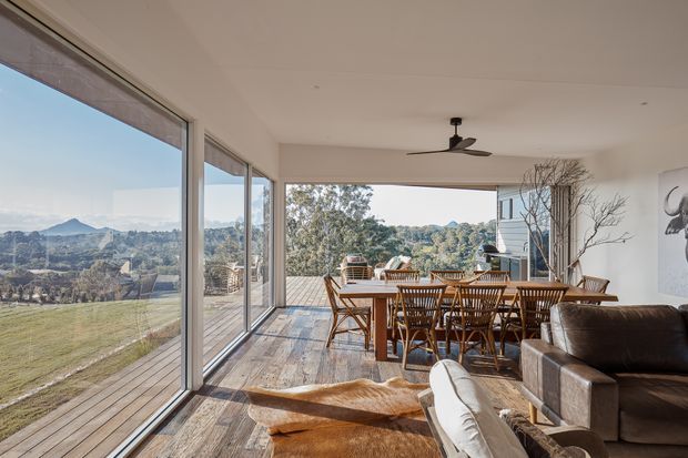A bright, spacious open-plan dining and living area with extensive floor-to-ceiling glazing overlooking a rural, hilly landscape. The room features a large timber dining table with woven timber chairs, timber flooring, a neutral sofa, a ceiling fan, and a deck extending beyond the open glazing. Natural daylight and warm timber tones create an inviting indoor-outdoor atmosphere.