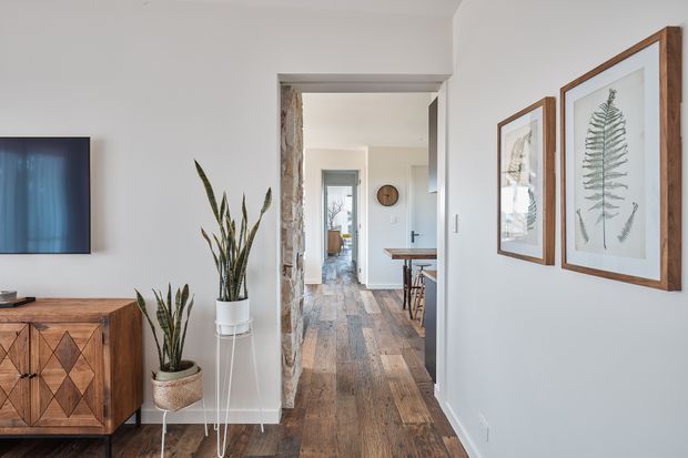 A bright residential hallway viewed from a living area, with white walls, wide-plank timber flooring, a timber console cabinet, potted plants, framed botanical artwork, and a wall-mounted television. The hallway leads toward a compact dining or kitchen area and a further glazed doorway. The interior has a warm, natural, contemporary character with mid-century-inspired timber furniture and understated decor.