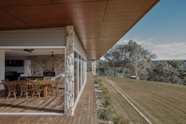 A covered timber deck and open-sided outdoor living area overlooking a sloping landscaped site. The space features a broad timber-lined soffit, timber decking, large sliding glass doors, and robust irregular stone-clad columns and fireplace walls. Inside, a dining table and chairs sit beside a stone fireplace, with a kitchen visible in the background. The setting is contemporary and strongly connected to the surrounding landscape.