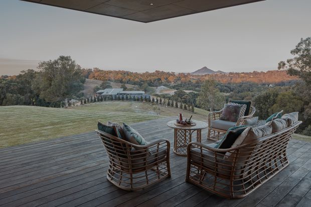 A covered outdoor terrace overlooking a landscaped rural valley, with a dark timber deck and a conversational seating arrangement of woven lounge chairs and a sofa around a small round table. The broad roof overhang creates a sheltered outdoor living space, while mature trees, lawns, distant hills, and autumn-toned vegetation form the background.