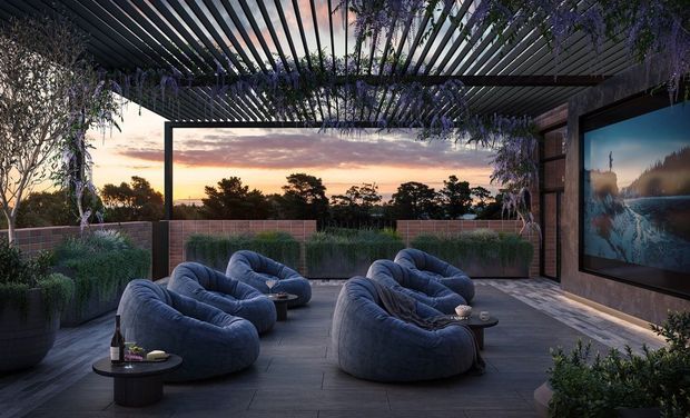 A sophisticated outdoor terrace lounge at sunset, featuring a slatted pergola overhead, lush climbing and potted greenery, dark tiled or stone paving, several deep blue lounge chairs, and a large wall-mounted artwork beside glazed or timber-clad walls. The terrace overlooks a planted landscape and distant sunset view, creating a refined, atmospheric entertaining space.