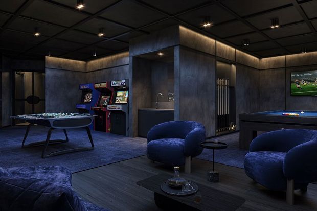 Dark, atmospheric entertainment room with charcoal textured walls, a black grid ceiling with recessed spotlights, deep blue carpet and furnishings, arcade machines, a foosball table, pool table, television, and lounge seating around a low coffee table. A compact bar or kitchenette with sink is built into the central partition.