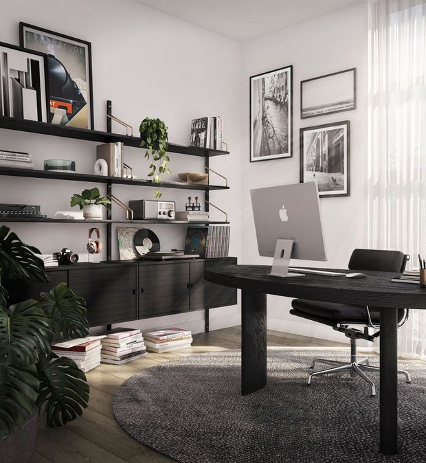 A refined home office with a dark timber desk, ergonomic black office chair, desktop computer, and extensive wall-mounted open shelving. The shelving displays books, artwork, audio equipment, records, and decorative objects, while large leafy plants add an organic element. Framed monochrome artwork hangs on the light wall, and sheer floor-to-ceiling curtains provide soft daylight. The room has dark timber flooring and a large textured grey rug.