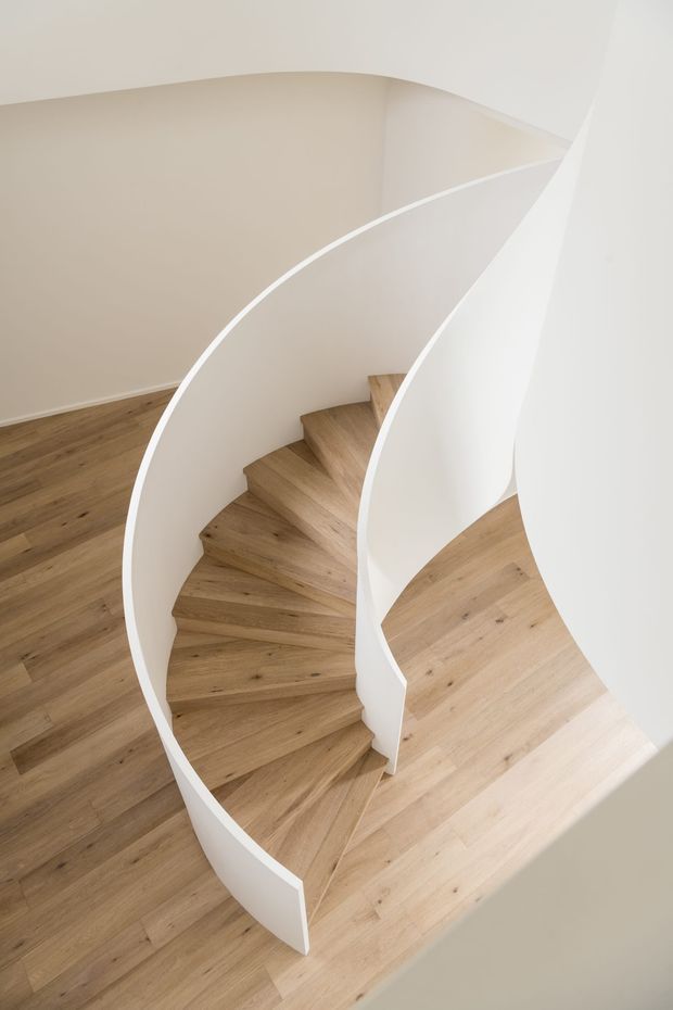 Interior view of a sculptural curved staircase with light timber treads and smooth white curved balustrade or walls. The staircase forms a sweeping spiral-like composition within a bright, minimalist space, with matching timber flooring and soft neutral walls.