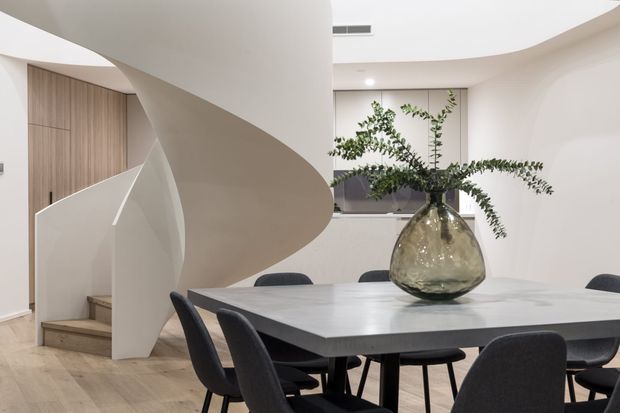 Contemporary open-plan dining and stair area with a sculptural curved staircase, pale timber flooring, smooth white walls and ceiling, a large dining table with dark upholstered chairs, and a substantial glass vase with greenery as the centrepiece. The space is bright, refined and minimalist, with concealed or integrated storage visible beside the stair.
