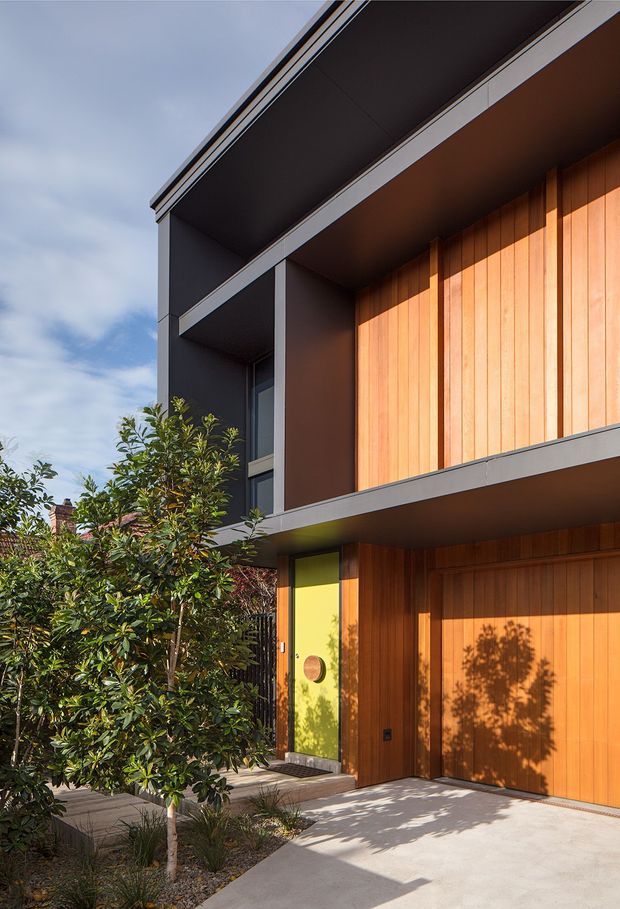 Street-level view of a contemporary multi-storey residential facade with a projecting upper balcony or canopy. The building combines dark charcoal cladding and trim with warm vertical timber wall cladding, a vivid lime-green entry door, and a large integrated timber garage door. A landscaped front garden and concrete driveway occupy the foreground.