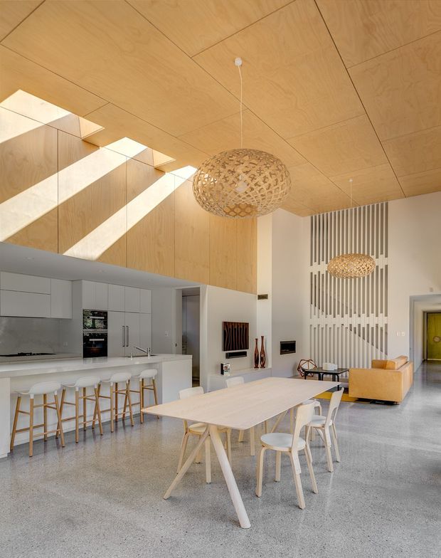 Bright open-plan kitchen, dining and living space with a high timber-lined ceiling, dramatic skylight openings, polished concrete floor, white kitchen cabinetry and island, pale timber dining table and chairs, and sculptural woven pendant lights. A vertical slatted feature wall defines the living area, which includes a tan sofa and contemporary artwork.