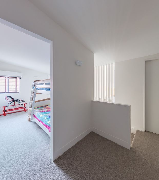 Bright residential hallway or landing with neutral painted walls, beige carpet, and a half-height balustrade beside a vertical-slat window. An open doorway reveals a children's bedroom with bunk beds and toys, while a closed interior door is visible on the right.