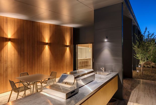 A sophisticated covered outdoor entertaining area with a built-in barbecue kitchen and dining setting. Warm vertical timber wall cladding is paired with dark metal wall panels, a pale stone benchtop, integrated stainless-steel barbecue appliances, and dramatic architectural lighting. The space opens toward a landscaped exterior and appears connected to an interior kitchen or dining area through glazing.