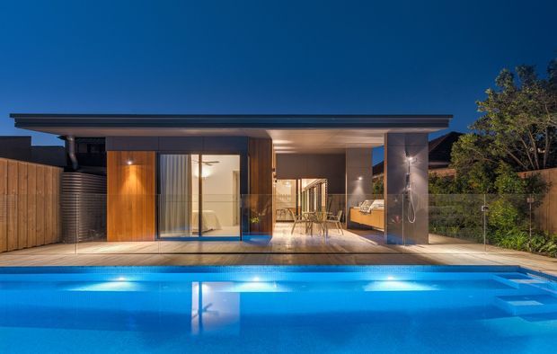 Twilight exterior view of a low, contemporary single-storey home opening onto a large illuminated swimming pool. The house has a broad flat roof with deep overhangs, extensive floor-to-ceiling glazing, warm timber wall cladding, dark framing, and a sheltered outdoor entertaining area. Interior spaces are visible through the glazing, including a kitchen or living area, while landscaped planting and timber fencing frame the property.