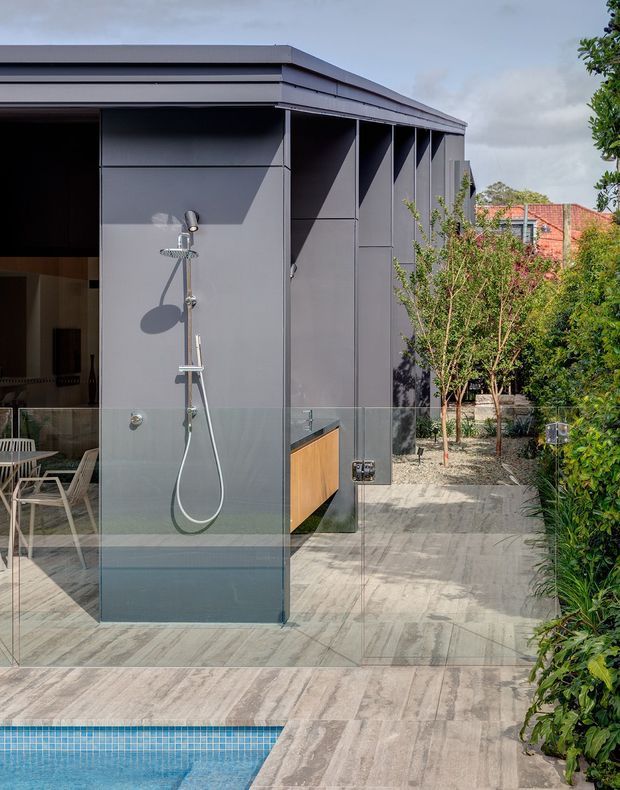 A contemporary outdoor poolside area featuring a frameless glass-enclosed shower with exposed chrome fixtures, set against a dark grey paneled building facade. Large-format pale stone-look paving surrounds the pool, with a timber-toned built-in cabinet or outdoor joinery visible behind the shower. Dense landscaped planting borders the walkway, and part of a dining area is visible through the glazing.