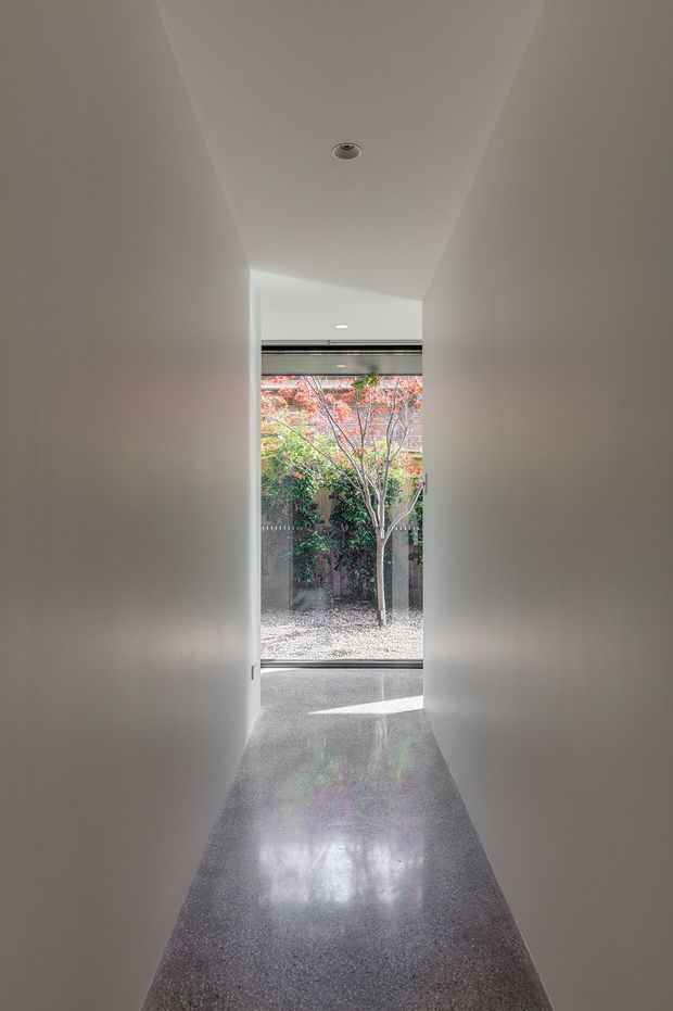 A narrow minimalist interior hallway with smooth white walls and ceiling, recessed downlighting, and a highly polished grey terrazzo or concrete-like floor. The corridor opens onto full-height glazing overlooking a small landscaped courtyard with a tree, bringing in natural light and creating a strong indoor–outdoor connection.