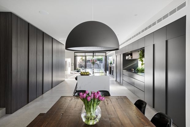 A sophisticated open-plan kitchen and dining area with a long dark timber dining table in the foreground, black pendant lighting, a white stone kitchen island, and extensive charcoal-grey built-in cabinetry. Full-height glazing at the rear opens the space to a bright courtyard or outdoor area, while integrated appliances, shelving, and greenery reinforce the clean contemporary design.