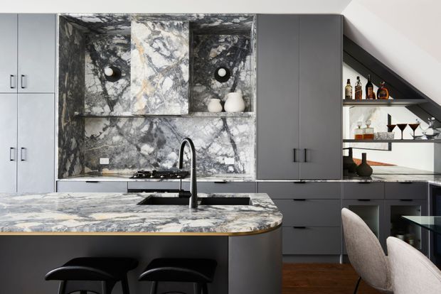 Contemporary kitchen with dark grey cabinetry, a substantial marble island and matching marble splashback, black sink and tapware, integrated shelving, and a compact drinks display beneath a sloping ceiling. Two stools sit at the island, with dining chairs visible in the foreground. The space has a refined, high-contrast material palette and built-in bar storage.