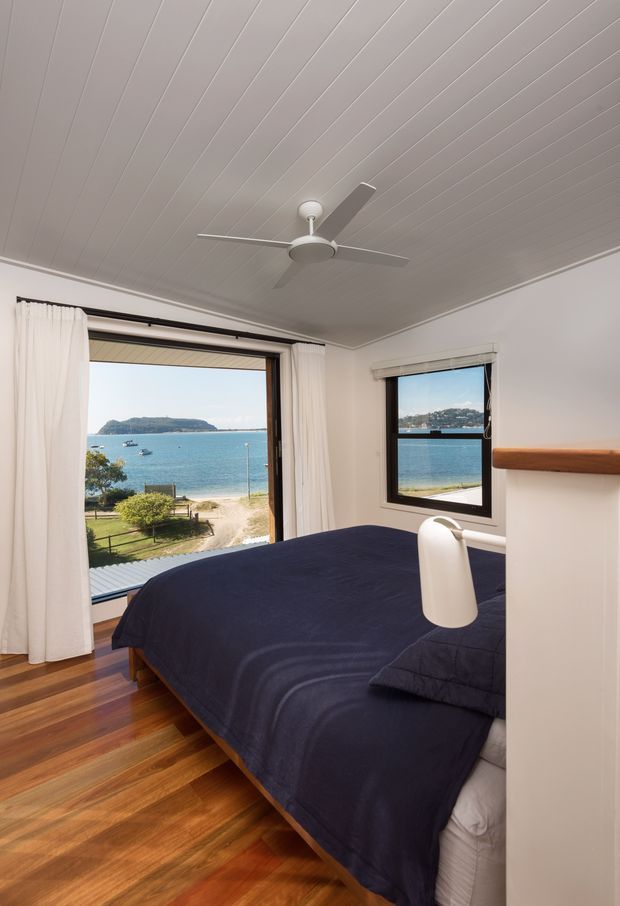 A bright coastal bedroom with a dark navy bed, white walls and curtains, warm timber flooring, and a ceiling fan. Large windows and a glazed opening provide direct views over a sandy beach, blue water, boats, and a headland. A simple timber wall shelf and pendant light add understated detail.