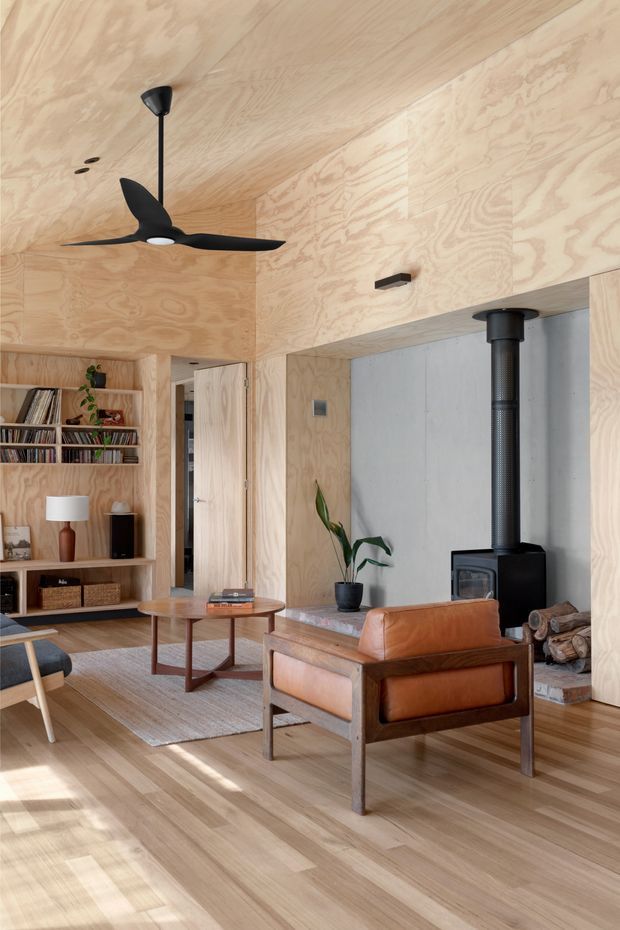 Warm contemporary living room with extensive pale plywood wall and ceiling lining, timber flooring, built-in open shelving, a black ceiling fan, and a freestanding black wood-burning stove set against a grey concrete or plaster feature wall. The room is furnished with a cognac leather lounge chair, low round timber coffee table, rug, and shelving with books and decor. Natural light creates a calm, refined atmosphere.