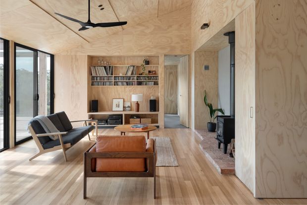 Warm, light-filled mid-century-inspired living room with extensive exposed plywood wall and ceiling linings, timber flooring, built-in open shelving and cabinetry, black-framed glazing, and leather lounge chairs arranged around a low coffee table. A compact black wood-burning stove is visible beside the built-in joinery, with a ceiling fan overhead and a neutral woven rug grounding the seating area.