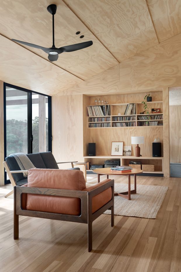 Warm, wood-lined living room with a mid-century-inspired leather armchair, dark sofa, round coffee table, built-in timber shelving filled with records and books, compact speakers, and a large black ceiling fan. Full-height glazing opens to a leafy outlook, while the plywood ceiling and walls create a cohesive natural-material interior.