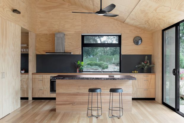 A warm, compact contemporary kitchen clad in natural plywood, with matching timber cabinetry, a dark stone island benchtop, black splashback and appliances, two bar stools, a ceiling fan, and large glazed doors opening to greenery outside.