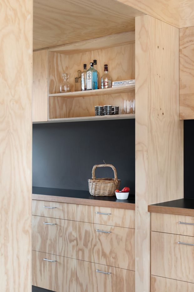 Close-up view of a custom plywood built-in bar or kitchen storage unit. Natural plywood cabinetry frames open shelves displaying bottles and glassware, with a dark charcoal backsplash and benchtop, lower drawers, and a wicker basket on the counter.