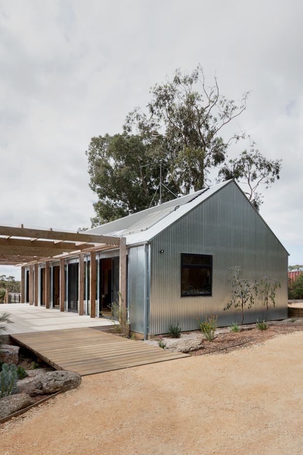 Exterior view of a compact single-storey dwelling with a steep gabled roof and corrugated metal cladding. A timber-framed pergola extends along the glazed frontage, connecting to a raised timber deck and boardwalk. The building sits in a dry, landscaped setting with gravel beds and sparse planting beneath an overcast sky.