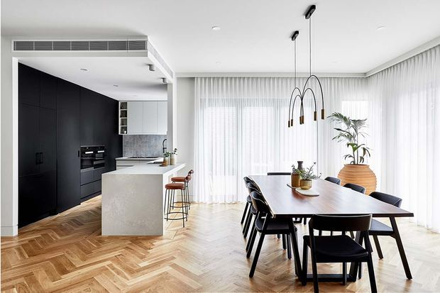 Bright open-plan kitchen and dining area with a large white island, matte black full-height kitchen cabinetry, integrated appliances, and a marble- or stone-look benchtop. The dining area features a dark timber rectangular table, black dining chairs, sculptural pendant lights, sheer floor-to-ceiling curtains, indoor plants, and a herringbone timber floor.