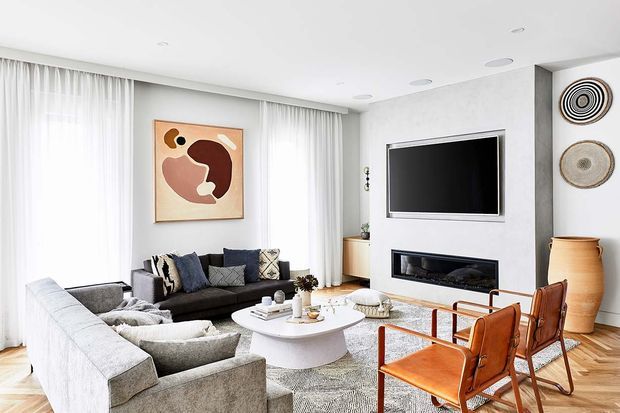 Bright contemporary living room with a large light-grey sectional sofa, dark sofa, round white coffee table, and two cognac leather sling chairs. Floor-to-ceiling sheer curtains frame the windows, while a TV and linear fireplace are integrated into a pale textured feature wall. Abstract artwork, woven wall decor, timber flooring, and neutral furnishings create a refined, warm interior.