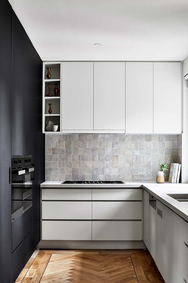 A compact contemporary kitchen with a U-shaped layout, matte white handleless cabinetry, a dark full-height wall housing integrated appliances, and a grey-beige textured tiled splashback. The kitchen includes a gas cooktop, corner sink area, open display shelving with decorative vessels, and a warm herringbone timber floor.