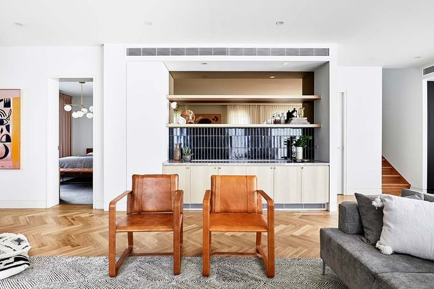 Bright open-plan living room viewed toward a built-in bar or kitchen joinery feature. The room has white walls and ceiling, warm herringbone timber flooring, a textured grey rug, two caramel leather armchairs, and a large grey sectional sofa. The built-in feature combines pale cabinetry, open timber shelving, a dark blue vertical tile splashback, and a high clerestory-style ventilation strip. A bedroom and stairway are visible through openings at the sides.