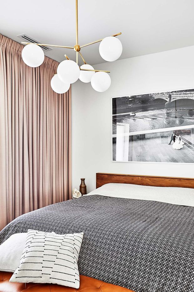 A stylish contemporary bedroom with a large bed, patterned charcoal-and-white bedding, a warm wooden headboard, and a rust-toned upholstered bed base. Full-height blush-pink curtains cover the window, while a sculptural brass-and-white globe pendant light hangs from the ceiling. A black-and-white framed photograph provides a graphic wall feature.