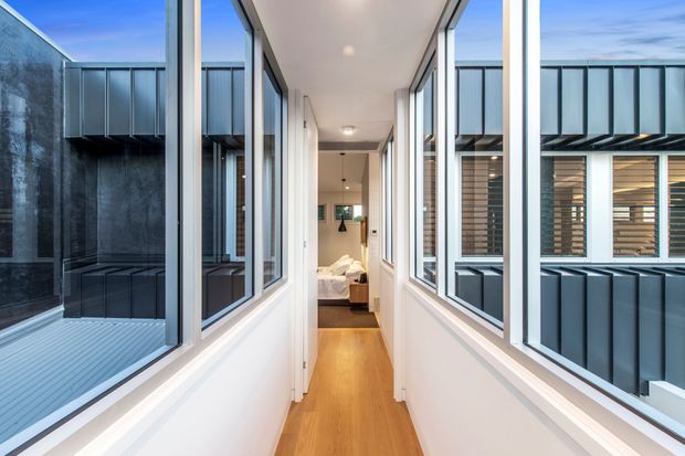 A bright, narrow interior corridor with white walls, recessed ceiling lights, and warm timber flooring. Full-height glazed openings on both sides overlook enclosed architectural courtyards or adjacent building volumes with dark metal cladding. A bedroom is visible at the far end of the hallway.