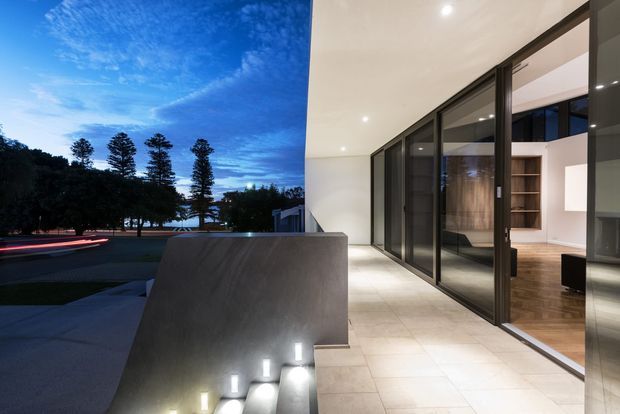 A contemporary indoor-outdoor transition space at dusk, featuring a broad tiled terrace or balcony beside full-height dark-framed sliding glass doors. The terrace overlooks landscaped grounds with tall trees and a distant waterfront-like horizon. A sculptural concrete or stone privacy wall incorporates illuminated steps leading down to the exterior landscape. Through the glazing, a warm timber-floored interior with built-in shelving is visible.
