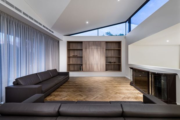 A refined contemporary living room with a large dark sectional sofa, herringbone timber flooring, full-height curtains, and a built-in timber wall shelving unit. A sleek dark fireplace or media joinery element sits on the right, while clerestory glazing and a high angled ceiling bring in natural light.