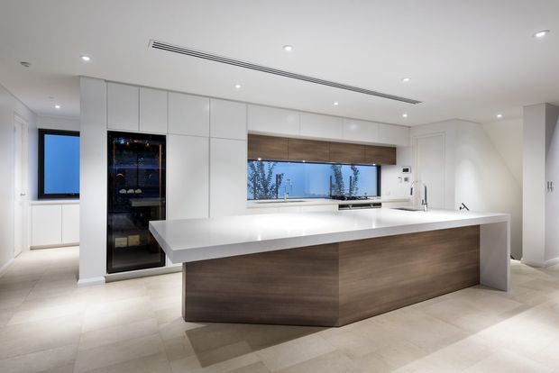 A bright, contemporary minimalist kitchen with a large white waterfall island, dark timber-look cabinetry, full-height white storage joinery, integrated appliances, and a wide horizontal window above the rear bench. Recessed ceiling lights and a linear air-conditioning vent contribute to the clean, streamlined fit-out.