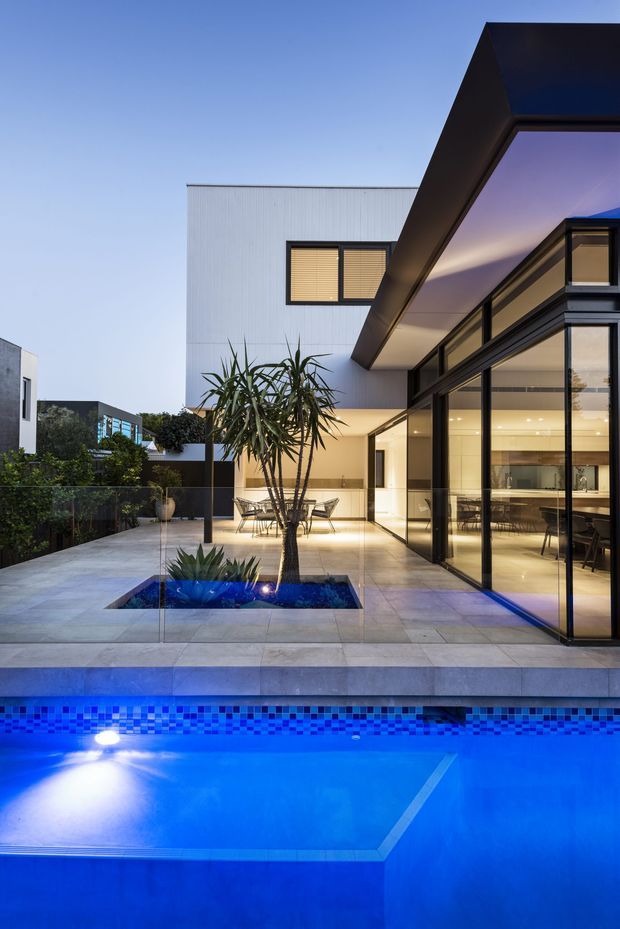 A dusk exterior view of a contemporary multi-level residence with a strong black-framed glazed corner and projecting angular roof canopy. The home opens onto a tiled terrace with a small planted water feature and a vividly illuminated swimming pool in the foreground. Warm interior lighting reveals a kitchen and dining area through the expansive glazing, while low garden planting and privacy screening surround the courtyard-like outdoor space.
