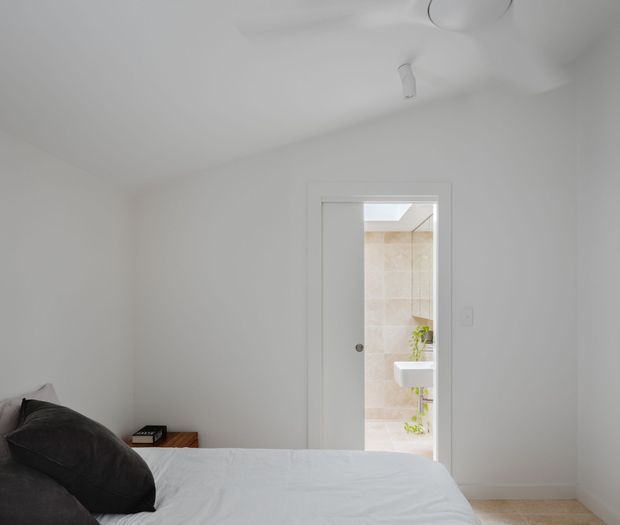 Bright, minimalist bedroom viewed from beside the bed, with white walls and ceiling, a low-profile bed dressed in white bedding, dark pillows, and a small timber bedside table. An open doorway leads directly to a light-tiled bathroom with a wall-mounted white basin and greenery. A skylight or roof window brightens the bathroom, while recessed and surface-mounted ceiling lighting provides additional illumination.