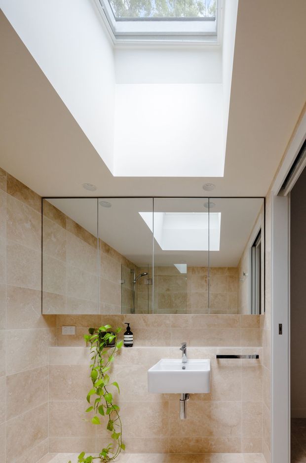 Bright contemporary bathroom with large beige stone-look wall and floor tiles, a wall-mounted white basin, chrome tap and exposed plumbing, frameless glass shower enclosure, mirrored cabinetry, and a large skylight that brings in natural light. A trailing plant adds a small organic accent.