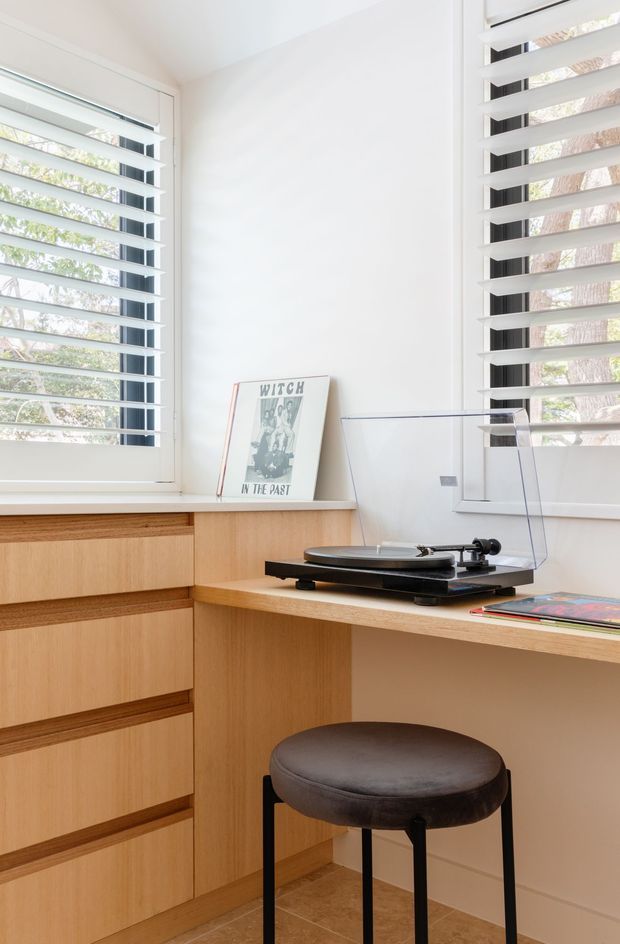 Bright, compact built-in timber study nook or home office with extensive drawer storage, a corner desk, turntable, framed artwork, and a round upholstered stool. White walls and plantation shutters bring in filtered natural light, while the warm timber joinery creates a clean, contemporary feel.