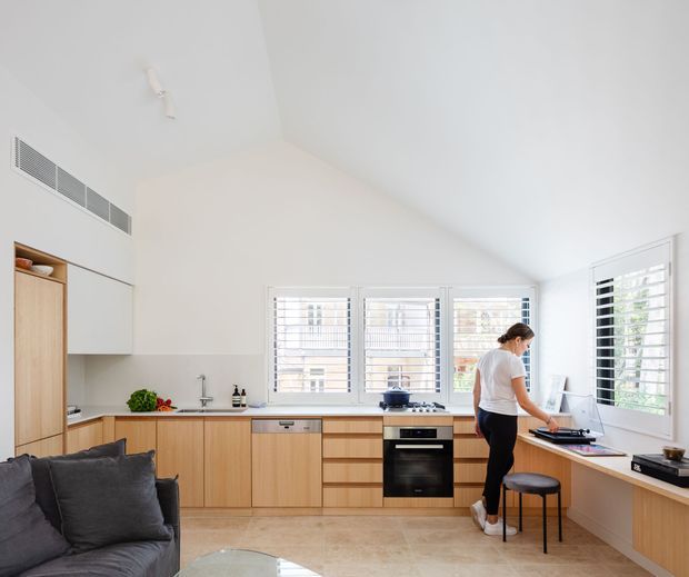 Bright open-plan kitchen with a high sloping ceiling, pale timber cabinetry, integrated appliances, white walls, and extensive plantation-shuttered windows. A peninsula or window-side work counter provides a small home workspace, while part of a dark sofa is visible in the foreground. The room has a clean, contemporary appearance with warm natural timber and abundant daylight.