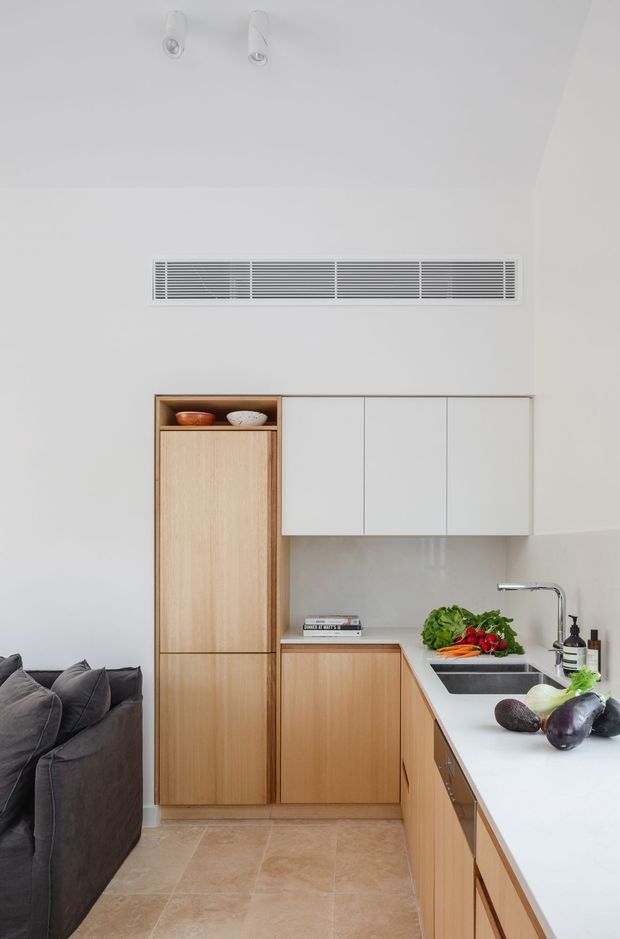 A compact, bright contemporary kitchen with light timber cabinetry, white upper cabinets, a long white benchtop, and an integrated double-bowl sink. A tall pantry cabinet includes an open display shelf, while fresh produce sits on the counter. A dark upholstered sofa is partially visible at the left, suggesting an open-plan living area. The room has a minimalist, clean-lined fit-out, pale stone-look tiled flooring, white walls, and discreet ceiling spotlights and ventilation.