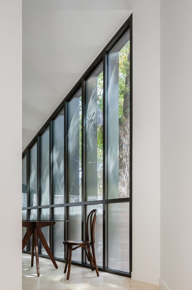 A bright, minimalist interior corner featuring a dramatic full-height wall of dark-framed glazing beneath a sharply sloped ceiling. A small timber table and curved wooden chair sit beside the windows, with trees visible outside. The space has pale walls, a light stone or polished floor, and a refined contemporary architectural character.