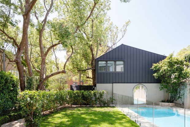 Exterior view of a contemporary residential pavilion set within a leafy landscaped garden. The dark vertically ribbed gable-ended upper facade contrasts with a light rendered lower level, which features an arched opening beside a bright blue swimming pool enclosed by frameless glass fencing. Mature trees, hedges and lawn surround the building.