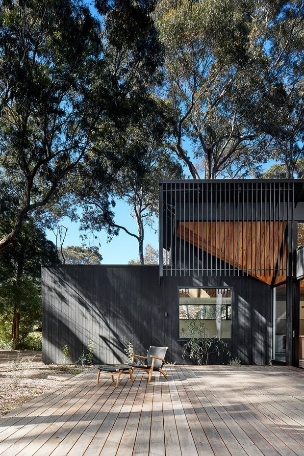 Exterior view of a modern dark-clad house set among mature trees. A broad timber deck occupies the foreground with two lounge chairs, while the facade combines dark vertical cladding or battens, large glazing, and a striking warm timber-lined upper section beneath a projecting roof.