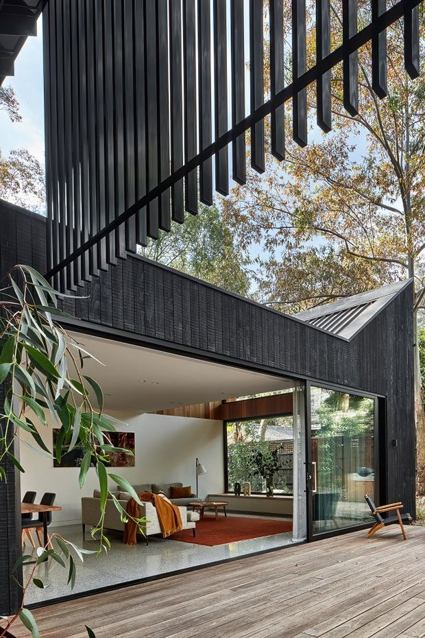 Exterior view of a dark, timber-clad modern home opening onto a broad timber deck. Large glazed sliding doors reveal a furnished living room, while prominent vertical fins and a projecting roof structure create a dramatic screened facade. Mature trees surround the house, and a small gabled roof volume is visible behind the facade.