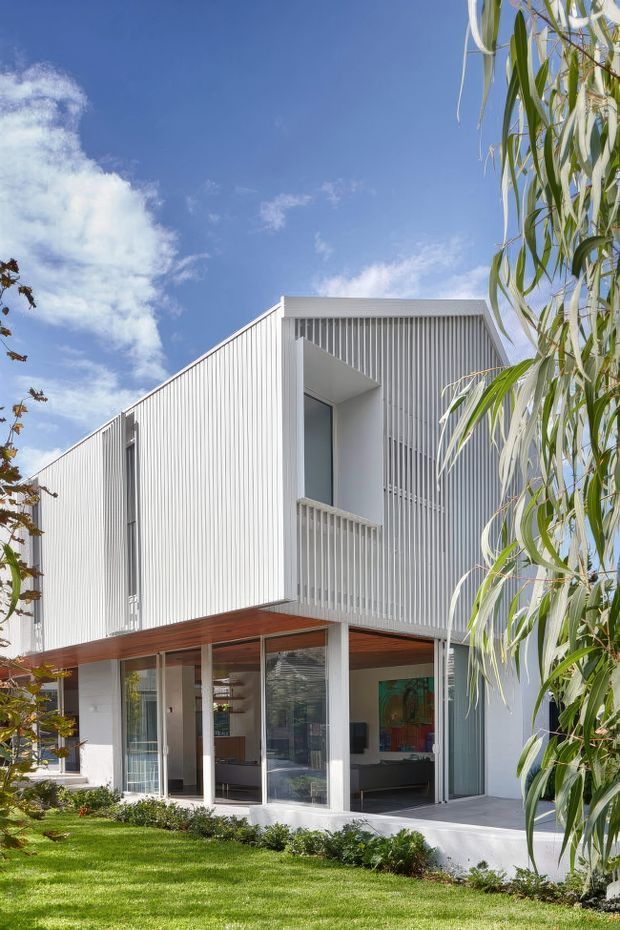 A bright modern two-storey residence viewed from the landscaped front garden. The house features crisp white vertically ribbed cladding, a projecting upper-level volume with a recessed balcony or opening, extensive ground-floor glazing, slender white columns, and a warm timber-lined soffit. Lawn, planting, and a tall leafy tree frame the facade under a clear blue sky.