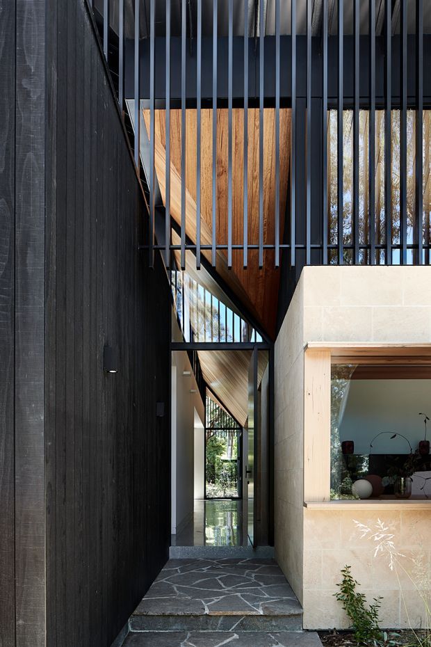 A narrow, dramatic architectural passage between a dark vertical timber-clad wall and a pale stone-clad volume. Black metal vertical screening creates a strong layered facade and reveals an upper-level timber-lined ceiling or soffit, while full-height glazing leads through the entry. Irregular dark stone paving, warm timber framing, and extensive reflections of surrounding trees create a refined contemporary atmosphere.