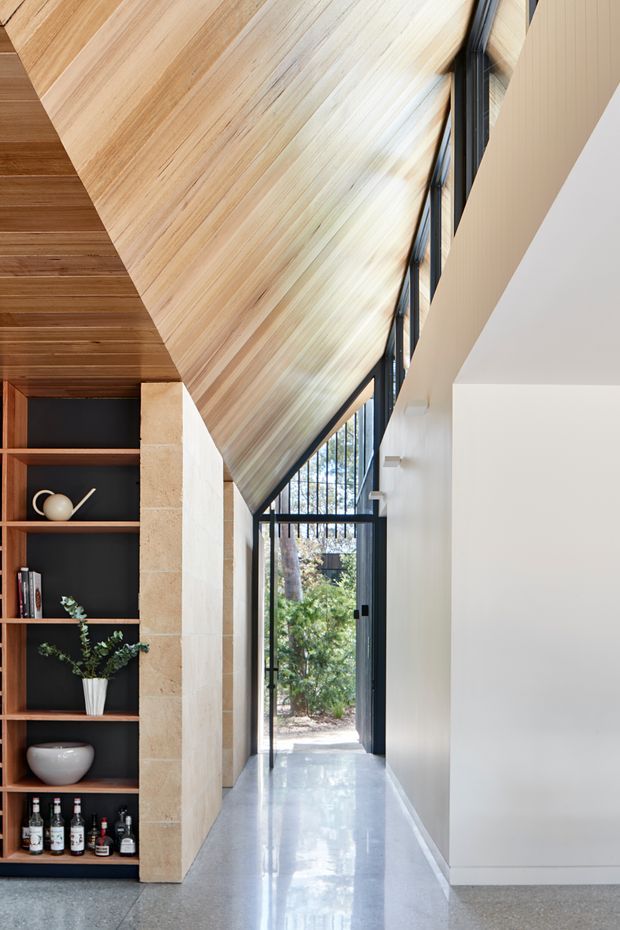 A narrow, refined interior hallway or entry passage with a dramatic sloping timber-lined ceiling, full-height black-framed glazing and a glazed exterior door at the end. A tall dark built-in shelving unit displays ceramics, books, a plant and bottles beside a pale stone-clad structural wall. The polished light-grey floor and white walls create a bright, contemporary architectural composition with strong vertical and diagonal lines.